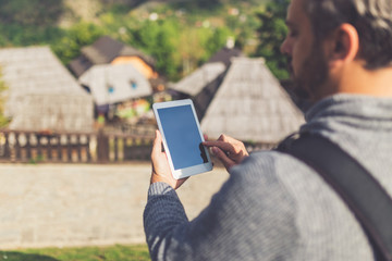 Rear view of young man using tablet device near retro mountain village wooden bungalows.