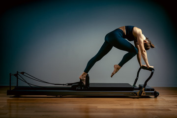 Young girl doing pilates exercises with a reformer bed. Beautiful slim fitness trainer on a reformer gray background, low key, art light, copy space advertising banner - obrazy, fototapety, plakaty