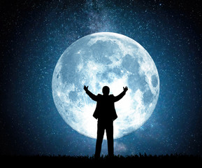 Silhouette of a man pulling his arms up on the background of the moon and the night starry sky.