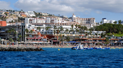 Naklejka premium Waterside distant view Los Cristianos coastline, people enjoy warm Atlantic Ocean pretty beach, touristic town, situated on south coast of Canary Islands, Spain