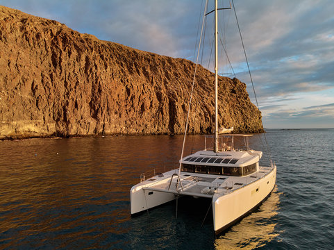 Moored lonely modern catamaran in calm waters of Atlantic Ocean near rocky volcanic cliff. Tenerife, Canary Islands, Spain. Concept of lifestyle, adventure activity, beautiful nature and freedom