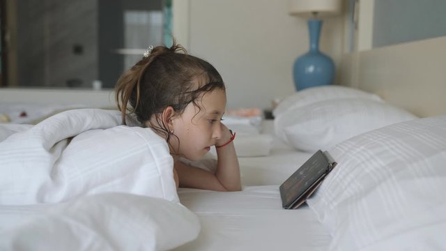 Little girl lying in bed under a blanket and watching cartoons on the tablet pc.