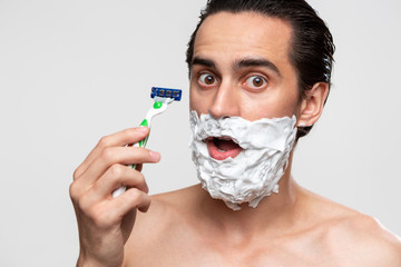 Handsome man with mustache uses shaving stick and shaving foam to trim his beard standning bare...