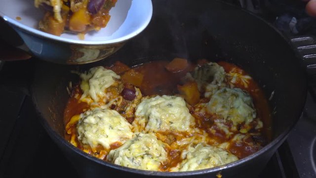 A slow motion close POV overhead shot of a man&rsquo;s hand lifting a ladle of steaming, cheese topped dumplings and vegetable stew, from a cast iron cooking pot on a stove hob, and serving it into a bowl..