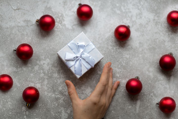 Woman's hand is taking a craft textured gift box with ribbon bow by unfocused vinous Christmas balls on a gray background, from above