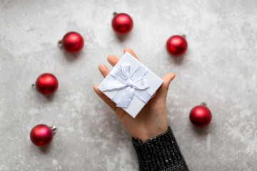 Woman's hand is holding a craft textured gift box with ribbon bow by unfocused vinous Christmas balls on a gray background, from above
