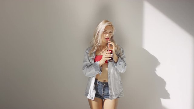 Thigh-up shot of lovely young woman in denim jacket, shorts and crop top gyrating provocatively against white wall, sipping red soda drink with straw and looking at camera with flirty gaze