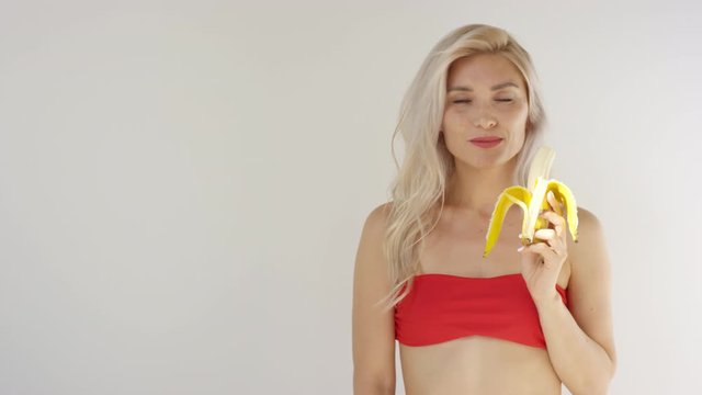 Waist-up shot of young blonde Caucasian woman in strapless crop top moving seductively, gazing at camera and chewing banana