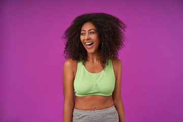 Portrait of beautiful young curly dark skinned woman in casual clothes posing over purple background with hands down, looking aside with joyful and charming smile, demonstrating white perfect teeth