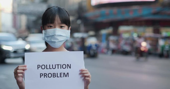 Little girl in protective mask holding pollution problem sign at city.