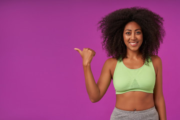 Positive young dark skinned woman with curly hair wearing sporty clothes, posing over purple background with raised hand and showing aside with thumb, looking at camera with happy and wide smile