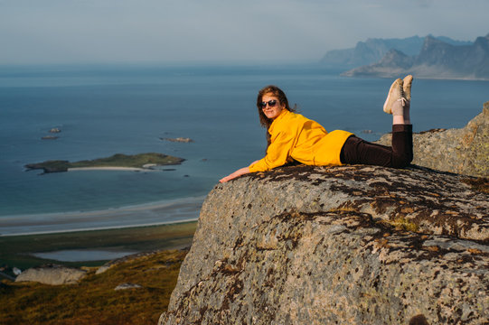 Smiling girl in yellow jacket and sunglasses lies on stone in on the edge of rocks in mountains and looking to camera