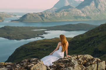 Back of girl in wedding dress sitting on rocks in mountains and looking to fjord - 299201832