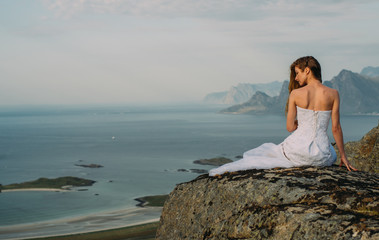 Fototapeta na wymiar Back of girl in wedding dress sitting on rocks in mountains and looking to fjord