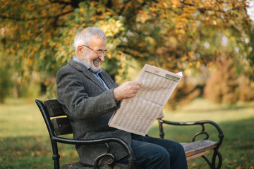 Bearded elderly man in glasses reading newspaper in the autumn park. Handsome gray-haired man sitting on the bench early in the morning