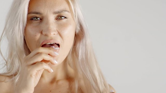 Close-up slow motion shot of young Caucasian blonde woman with bare shoulders putting two tabs of gum on tongue in mouth and chewing, while staring sensually
