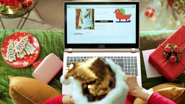 Holiday season. Upper view of young housewife in sparkle gold sequin santa hat sitting on divan in the modern living room at Christmas browsing high fashion retail online store on a laptop.