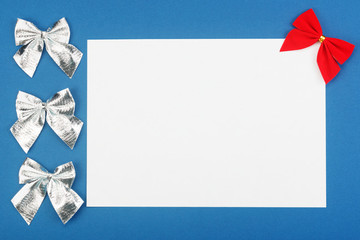 Blank white paper with red and silver christmas bows on blue background. Holidays mock-up with copy space for your greeting text. Flat layout.