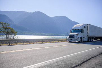 Big rig white semi truck with grille guard transport cargo in refrigerated semi trailer running on the road along river with mountain on the background