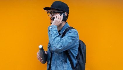 Portrait of young self-confident hipster man on orange background. Calling by smartphone with steel thermo eco bottle for water in another hand.