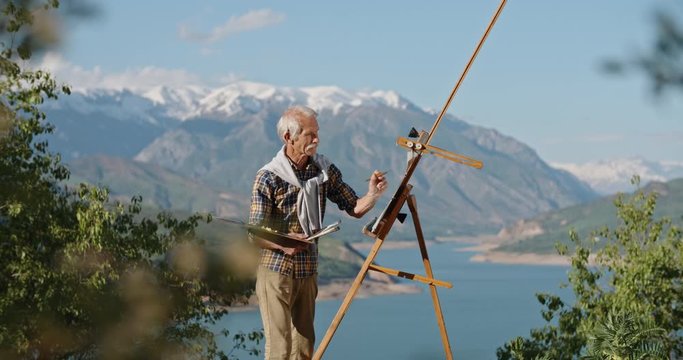 Retired caucasian man standing near easel, drawing a picture, enjoying his hobby on pension, looking for inspiration in beautiful landscape 4k