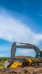 Vertical frame Excavator at a construction site with mountain homes and cloudy sky background
