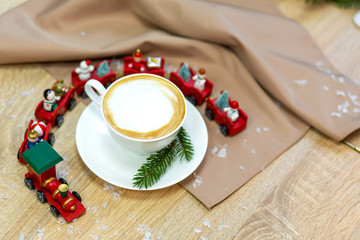 Delicious fresh festive morning cappuccino coffee in a ceramic white cup on the wooden table with decorative christmas train, red ornamentals, fireflies and spruce branches