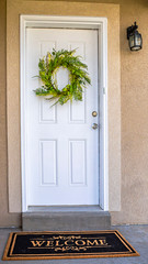 Vertical frame White front door with green leafy wreath and doormat at the facade of a home