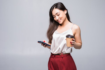 Portrait of a smiling asian woman using mobile phone while holding cup of coffee to go and looking away isolated over white background