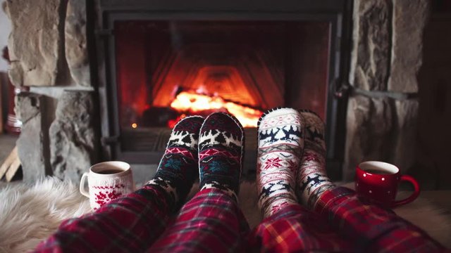 Feet in woollen socks by the Burning Christmas Cozy Fireplace. 4K. Couple relaxes by warm fire with cup of hot drink and warming up their feet. Winter and Christmas holidays concept.