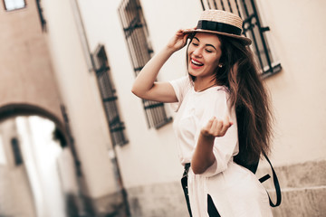 Young stylish woman walking on the old town street, travel with backpack, straw hat, wearing trendy outfit.