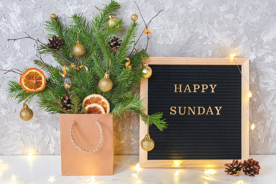 Happy Sunday text on black letter board and festive bouquet of fir branches with christmas decor in craft package and light lamp garland on table. Template for postcard Concept Hello winter Sunday