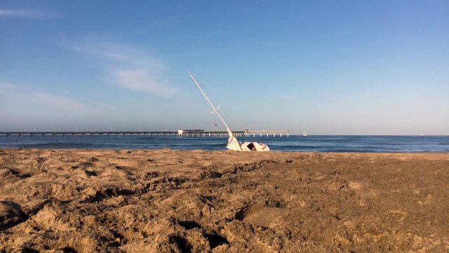 A shipwrecked sail boat washed ashore after a storm. Broken sails, worn flag, the boat is shoved around by the shore break.