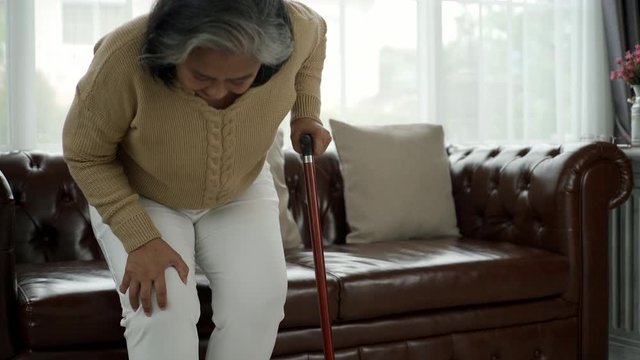 Senior adult with a painful expression from a serious legs and knee pain having difficulty getting up, sitting down on the sofa and moving around the house