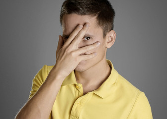 The man covers his face with his hand and peeks through his fingers. In a yellow t-shirt on a gray background