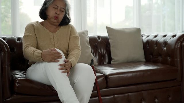 Senior adult with a painful expression from a serious legs and knee pain having difficulty getting up, sitting down on the sofa and moving around the house