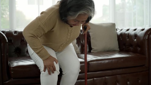 Senior adult with a painful expression from a serious legs and knee pain having difficulty getting up, sitting down on the sofa and moving around the house