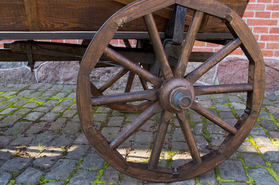 A brown wheel with a wooden rim and spokes at the left rear and the bottom of an vintage cart standing on an old gray stone sidewalk overgrown with grass against a red brick wall