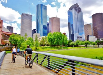 Bicyclists cross wooden bridge in Buffalo Bayou Park, with a beautiful view of downtown Houston...