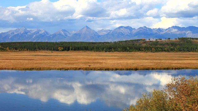 Beautiful autumn calm landscape with reflection in the river of a blue sky with clouds and a mountain range of the East Sayan. Natural autumnal background. Siberia, Buryatia, Tunka Valley