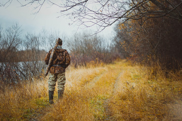 A man in camouflage clothing with a gun in a case on his shoulder, standing back on the forest road. Autumn hunting.
