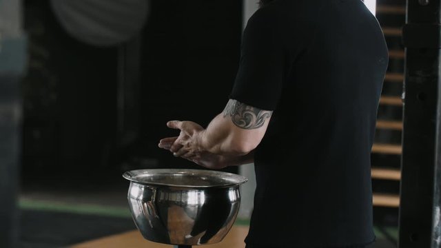 Close-up back view male weight lifter athlete applying talc powder before exercising in large hardcore gym hall.