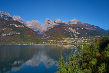 Aerial view of Lake Molveno, north of Italy in the background the city of Molveno, trees, Alps, blue sky. Reflection of mountains in water. Autumn season. Multi-colored palette of colors