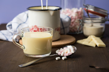 Zubereitung von heißer weiße Schokolade im Milchtopf mit Kakao Pulver und Marshmallows in Küche auf Holz Arbeitsfläche dunkel und Lila Hintergrund