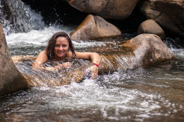 A young girl with dark hair smiles while sitting in the water of a waterfall. Large brown granite stones form a threshold for water with a natural pool.