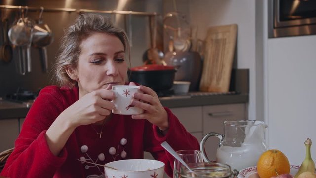 Young woman eating a delicious healthy breakfast with yoghurt in her kitchen surrounded by Christmas decorations and festive cutlery taking a big sip from her hot tea