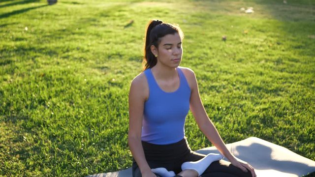 A fit young woman sitting in lotus position meditating in the park at sunrise to improve her mental health and relax.