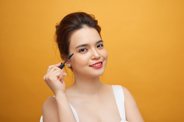 Closeup portrait of attractive young woman putting some mascara onto her eyelashes with make up brush over yellow background