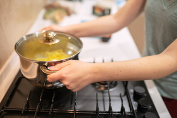 Beautiful young woman housewife prepairing dinner, hold in hands big steel saucepan, standing it on gas-stove.