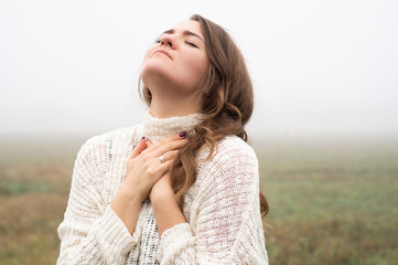 Girl closed her eyes, praying in a field during beautiful fog. Hands folded in prayer concept for...